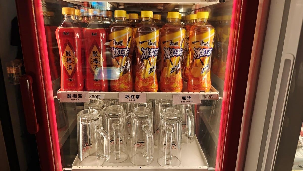 Bottled drinks in a fridge with glass mugs below, price tags visible.