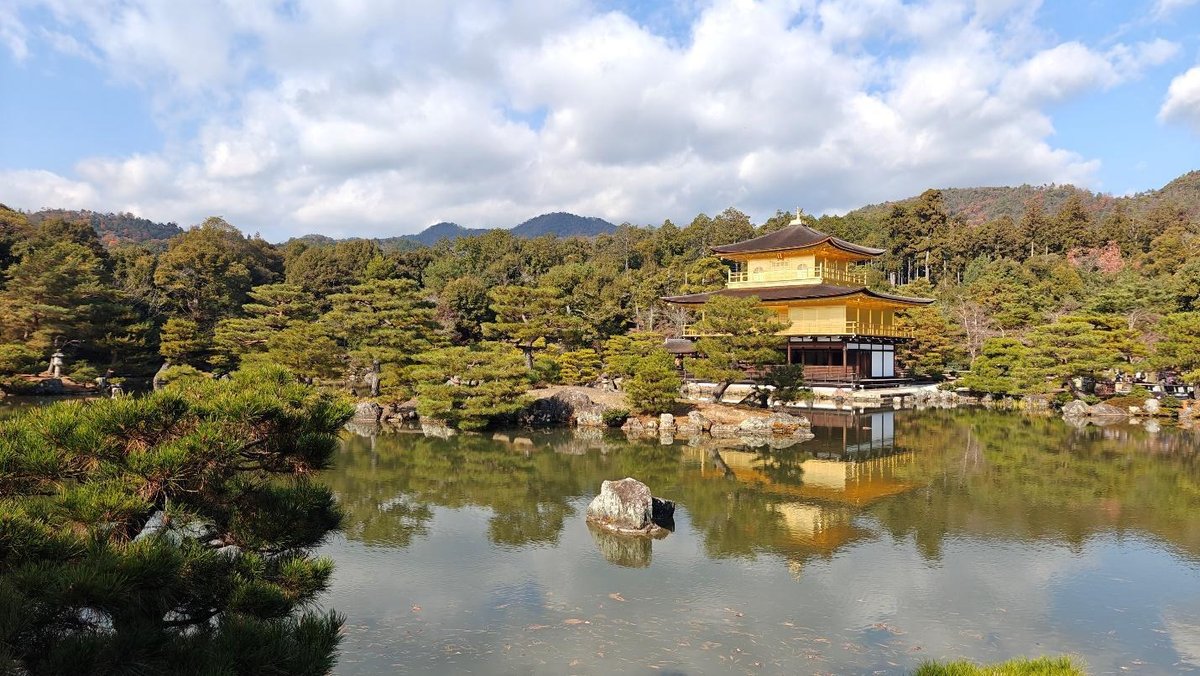 Golden pavilion by tranquil pond, surrounded by lush greenery and clouds.