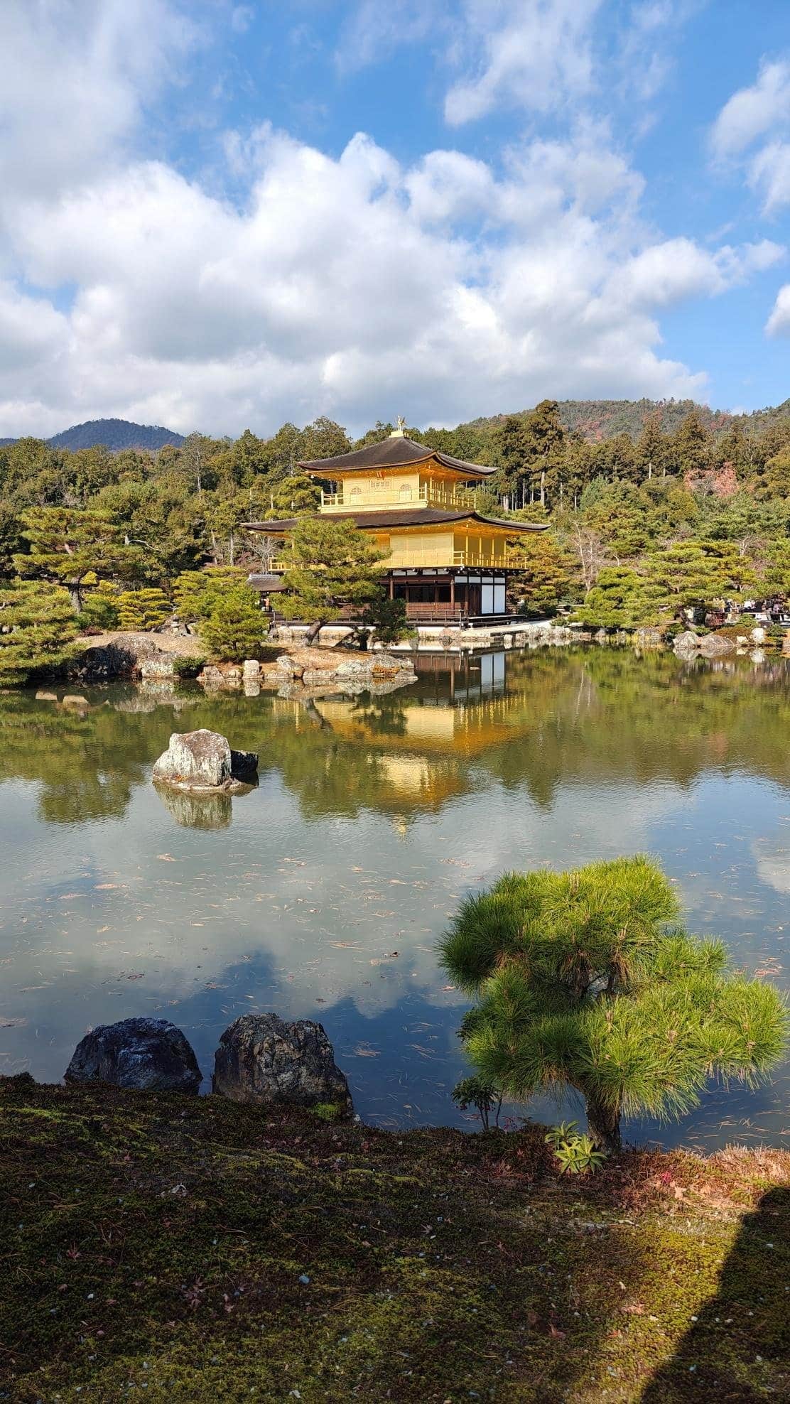 Golden pavilion reflecting in serene pond under blue sky and clouds.