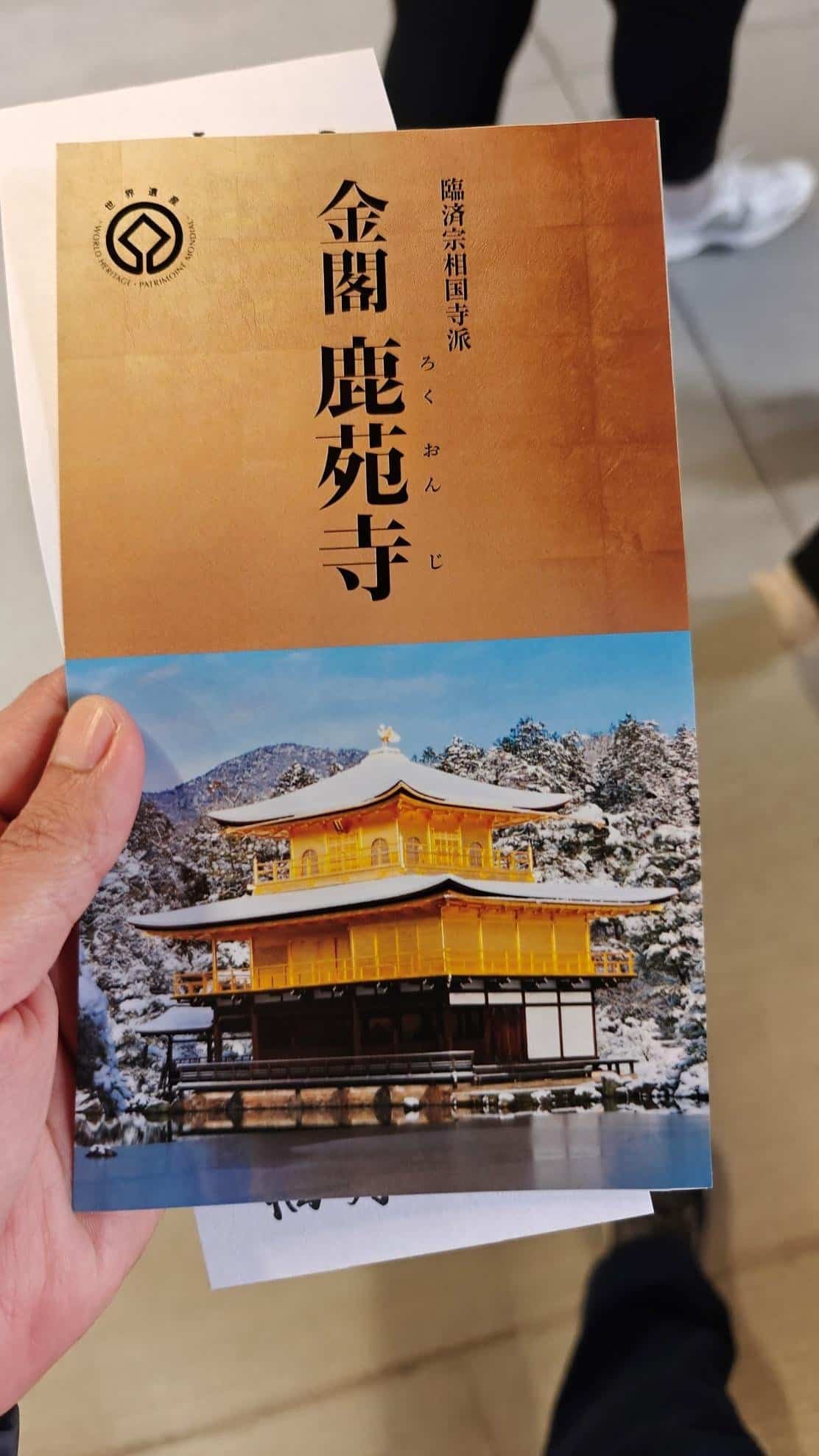 Hand holding brochure of Kinkaku-ji temple, snowy backdrop, gold facade.