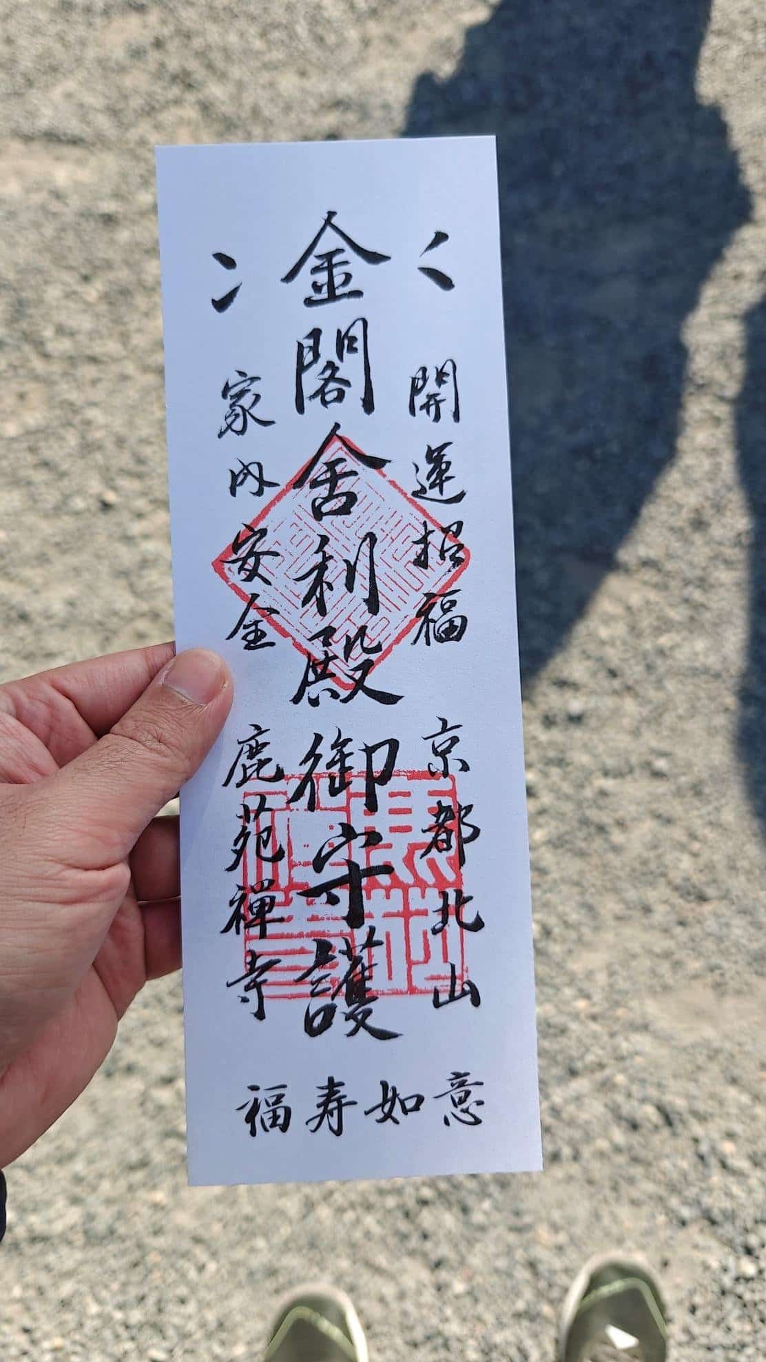 Hand holding Japanese prayer paper with text and red stamps, outdoor setting.