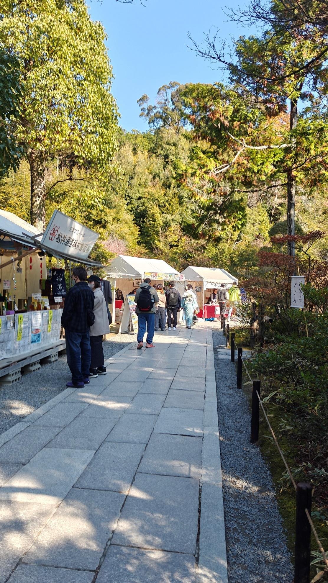 Path lined with market stalls, people, and lush trees under a clear sky.
