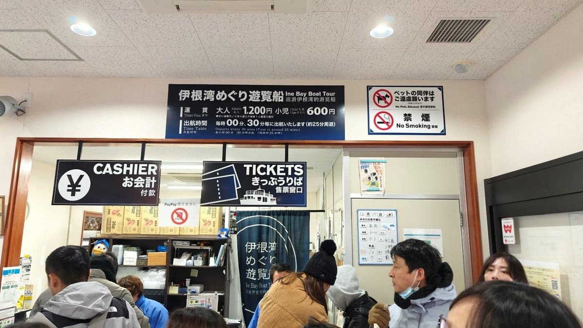 People queuing at a ticket and cashier counter, Ine Bay Boat Tour signs above.