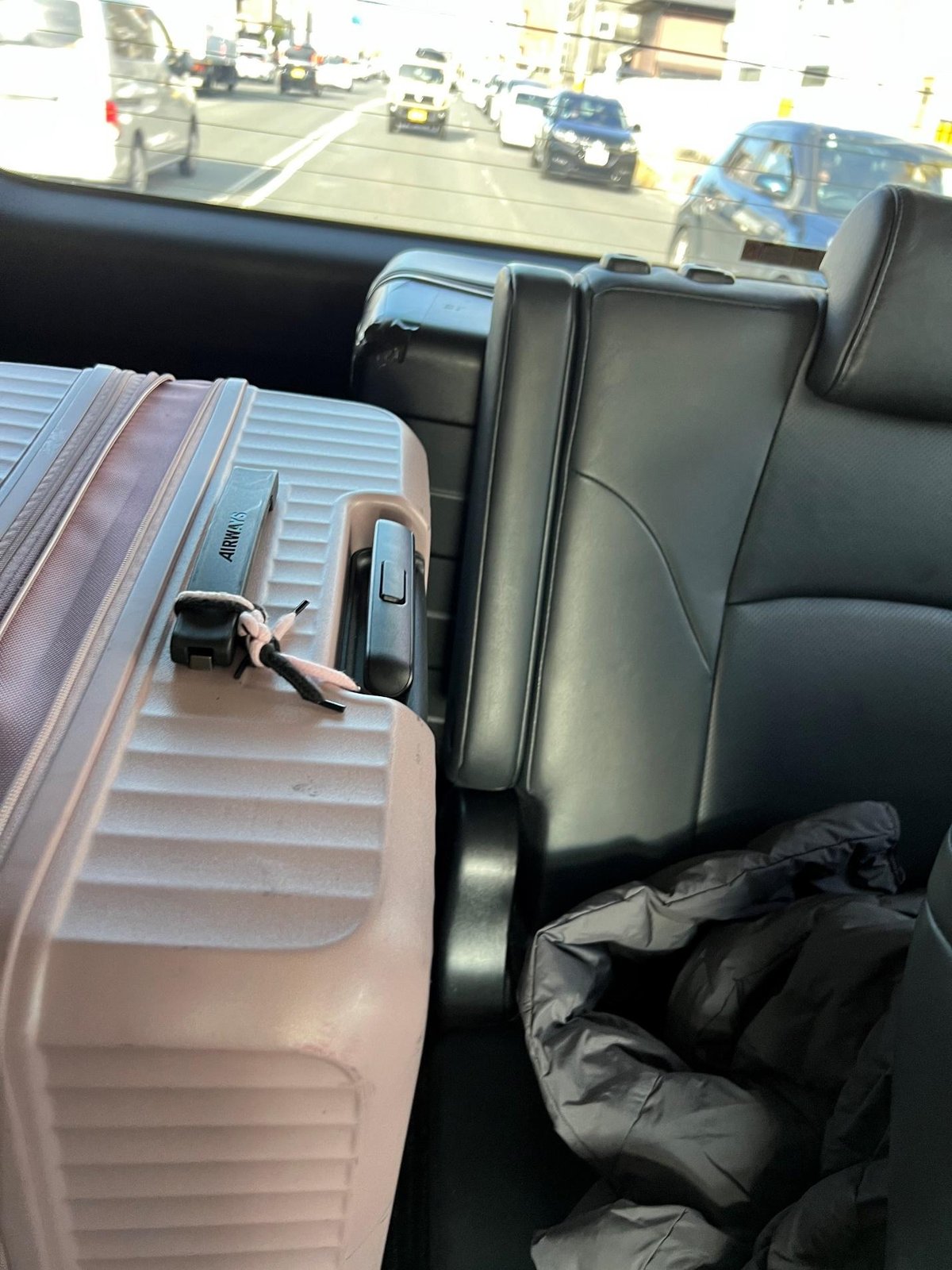Pink suitcase and gray jacket on black leather car seat, traffic view.