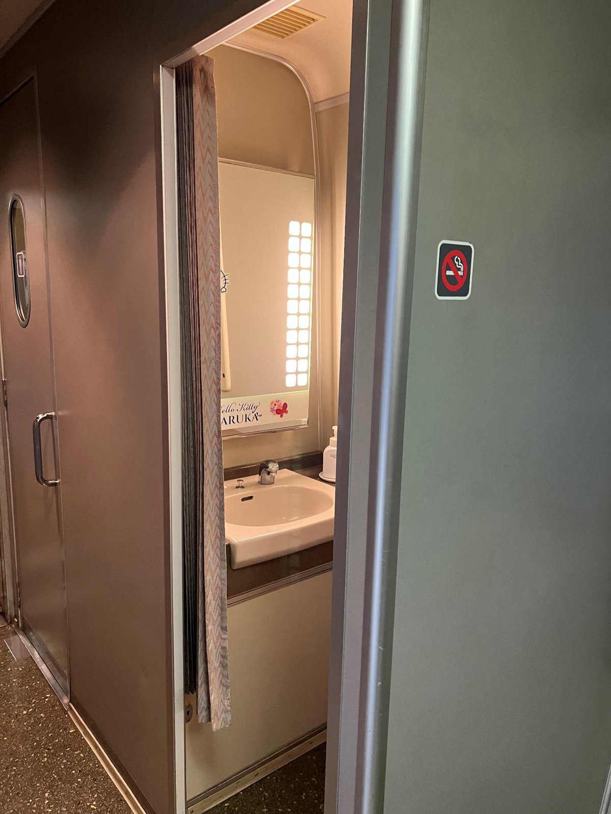 Train bathroom with sink, mirror, curtain, and no smoking sign.