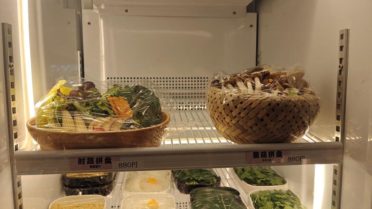 Vegetables and mushrooms in baskets, wrapped in plastic on a fridge shelf.