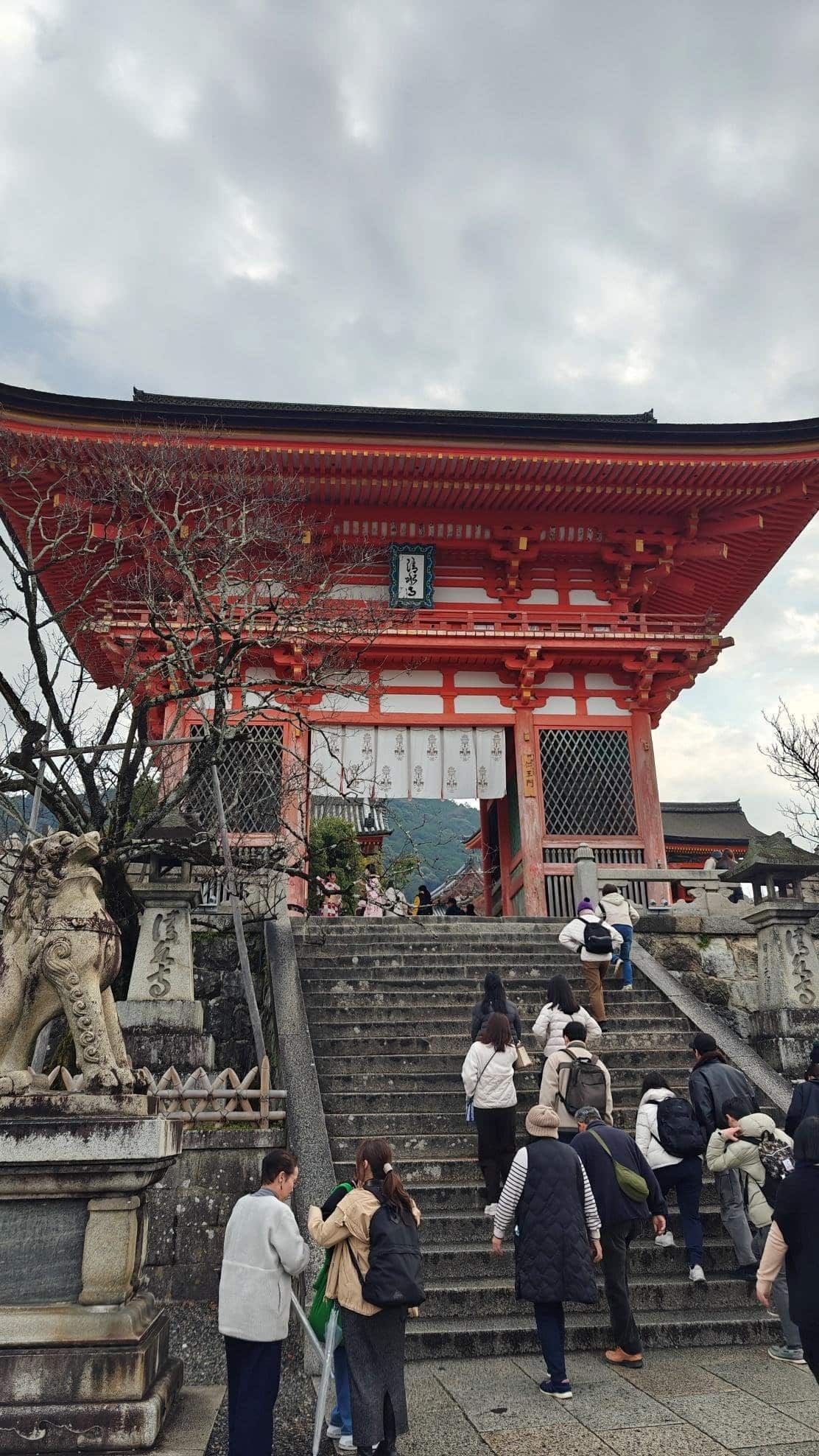 People ascending steps to large red temple gate under cloudy sky