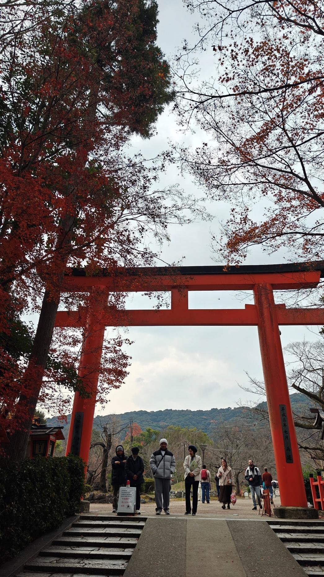 People beneath red torii gate with autumn trees, cloudy sky
