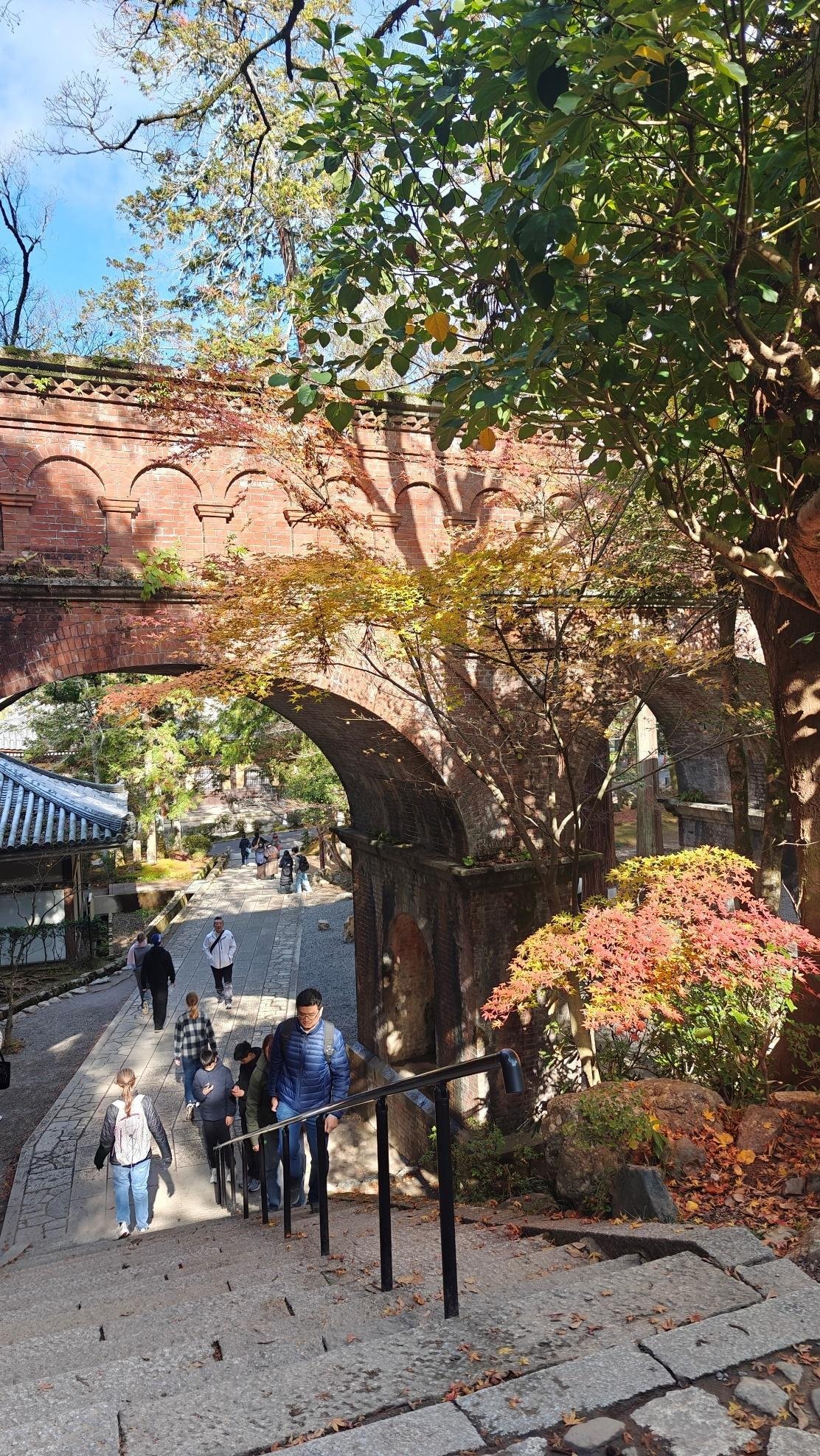 People climbing steps near a red brick arch and autumn trees.