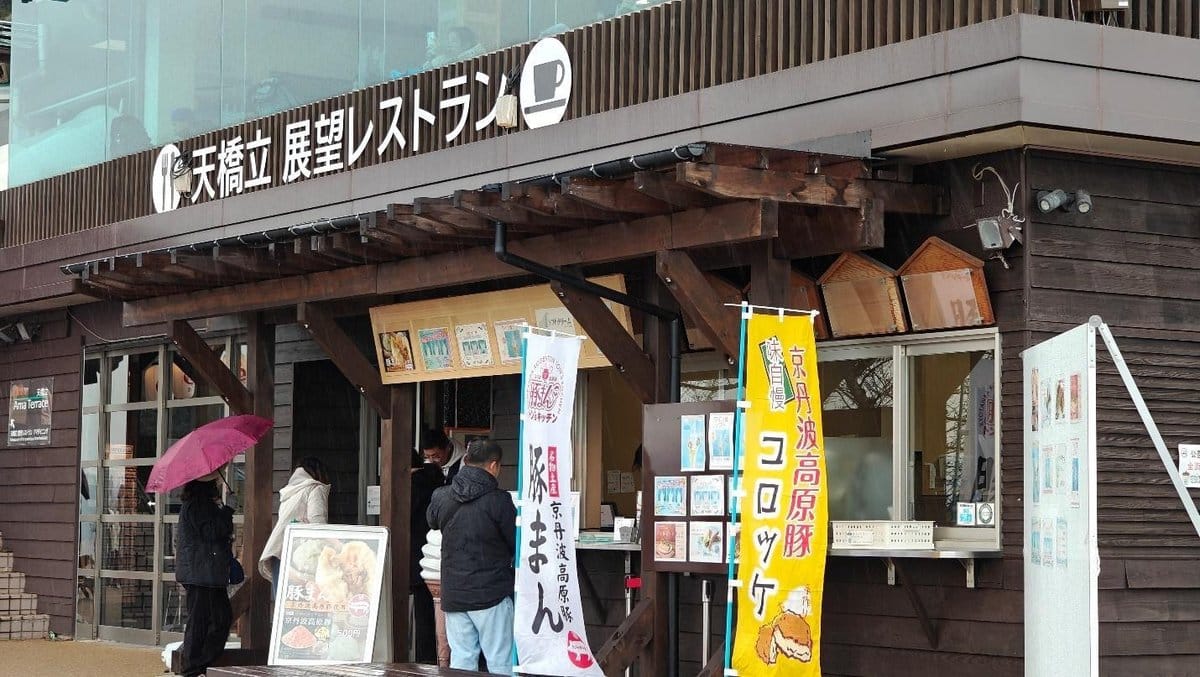 People with umbrellas outside a wooden cafe with colorful banners.